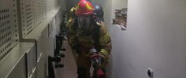 Zdjęcie przedstawia strażaka, który znajduje się w archiwum sądu. Posiada ubranie specjalne, ma założony aparat powietrzny oraz maskę i trzyma w ręku prądownicę wodną. 