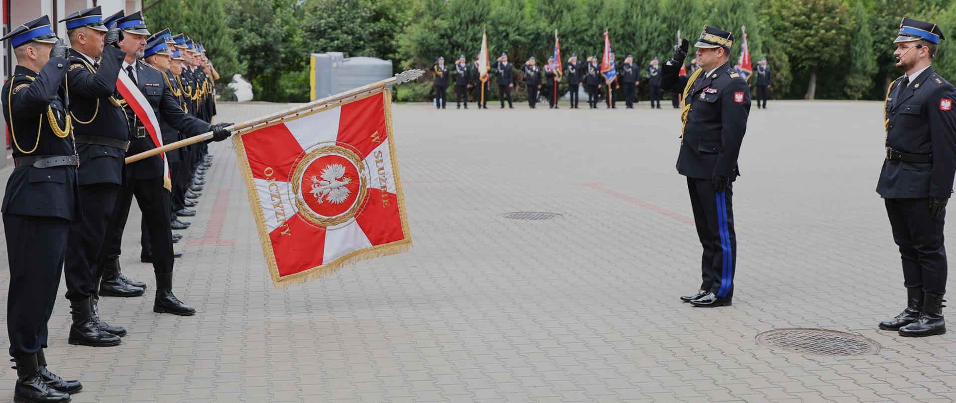 Podczas uroczystego apelu funkcjonariusze w mundurach stoją w szeregu i salutują sztandarowi. Na środku placu Mazowiecki Komendant Wojewódzki Państwowej Straży Pożarnej oraz dowódca uroczystości oddaje honor przed pocztem sztandarowym, który prezentuje czerwono-biały sztandar z godłem Polski. W tle widoczne są kolejne poczty sztandarowe oraz zadrzewiony teren otaczający miejsce ceremonii