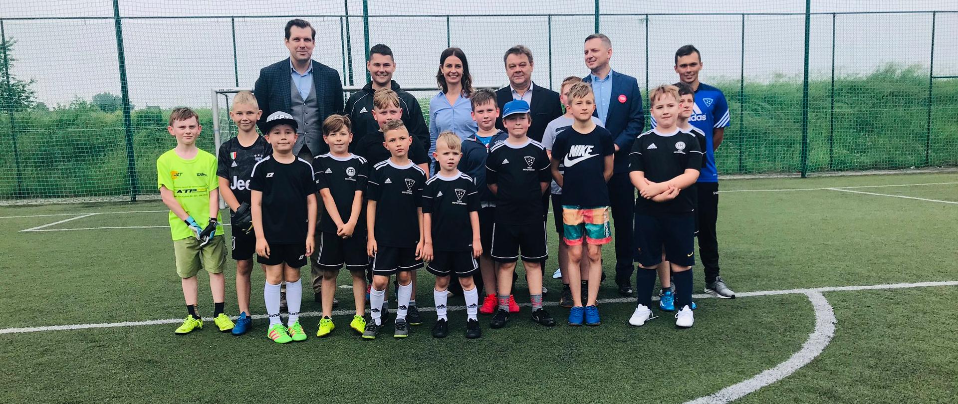 Minister Sportu Danuta Dmowska-Andrzejuk na grupowym zdjęciu na boisku piłkarskim.