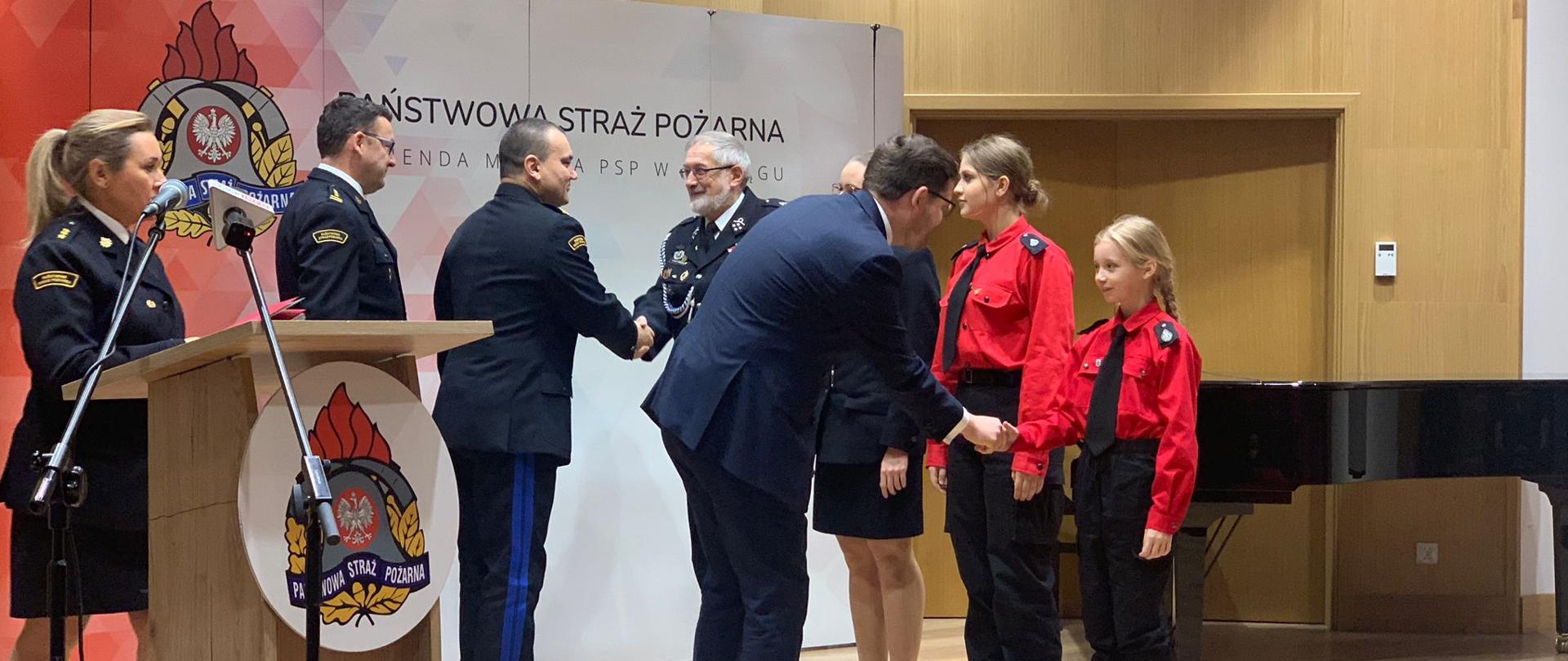 Przedstawiciele Państwowej Straży Pożarnej oraz Wojewoda Warmińsko Mazurski podczas wręczania promes przedstawicielom młodzieżowych drużyn pożarniczych. W tle czerwony baner z logiem państwowej Straży Pożarnej.