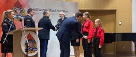 Przedstawiciele Państwowej Straży Pożarnej oraz Wojewoda Warmińsko Mazurski podczas wręczania promes przedstawicielom młodzieżowych drużyn pożarniczych. W tle czerwony baner z logiem państwowej Straży Pożarnej.