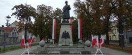 Uczczenie rocznicy. Plac przed pomnikiem Żołnierza Polskiego w Rawiczu. W centralnym punkcie pomnik. Na cokole postać Żołnierza Polskiego trzymająca broń. Na frontowej ścianie cokołu zawieszony orzeł w koronie. Za pomnikiem znajdują się cztery maszty, na których zawieszone są flagi państwowe. Z przodu pomnika stoją dwa stojaki z flagami państwowymi oraz trzy tablice pamiątkowe. W tle drzewa, budynki, przechodnie i ruch uliczny.