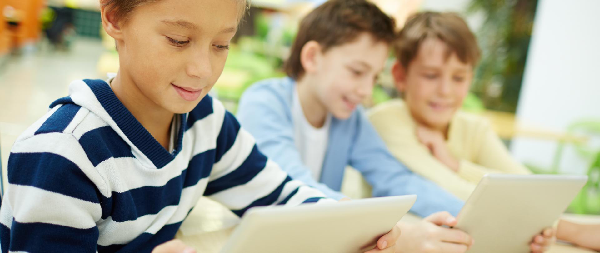 Zdjęcie w poziomie. Trzech chłopców siedzi przy stole. Korzystają z tabletów.