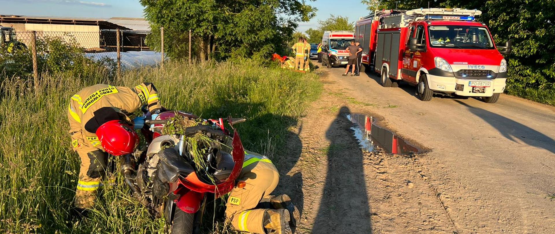 Na zdjęciu strażacy przy motocyklu na jezdni samochód ratownictwa technicznego a za nim karetka pogotowia