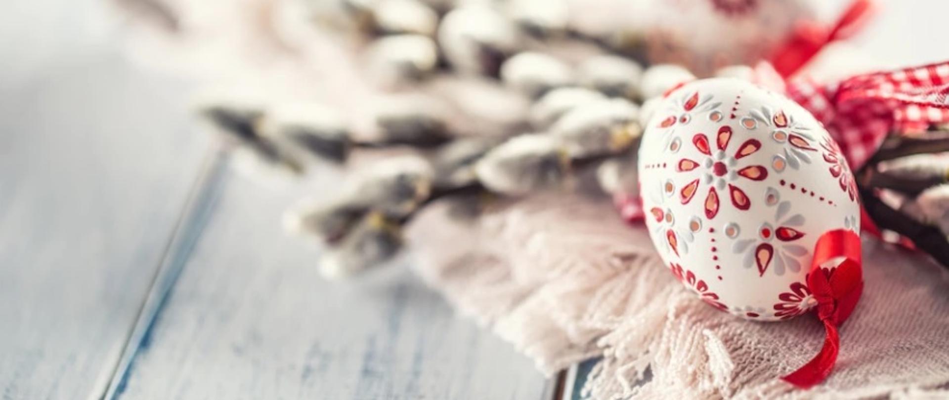 Grafika przedstawia pisankę z czerwoną wstążką, położoną na obrusie. W tle wielkanocne bazie.