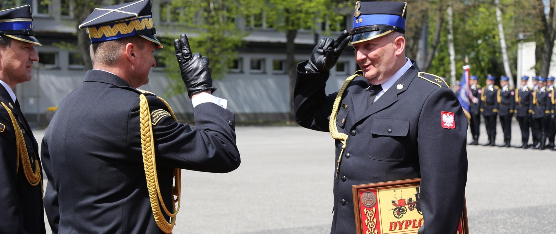 Zastępca Komendanta Głównego PSP wraz z komendantem szkoły stoją naprzeciwko strażaka, który trzyma w dłoniach otrzymany dyplom komendanta głównego PSP, Zastępca Komendanta Głównego PSP i funkcjonariusz salutują