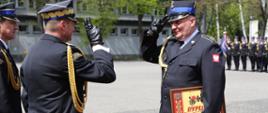 Zastępca Komendanta Głównego PSP wraz z komendantem szkoły stoją naprzeciwko strażaka, który trzyma w dłoniach otrzymany dyplom komendanta głównego PSP, Zastępca Komendanta Głównego PSP i funkcjonariusz salutują