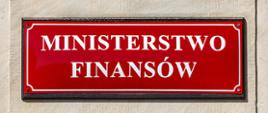 Czerwona tablica z napisem „MINISTERSTWO FINANSÓW” zamocowana na jasnej ścianie budynku.