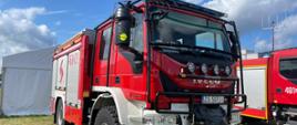 Zdjęcie przedstawia terenowy pojazd pożarniczy marki Iveco na co dzień służący strażakom podczas pożarów lasów. W tle biały namiot oraz lekko zachmurzone niebo. Na zdjęciu widać dodatkowo bok innego pojazdu oraz maszt antenowy.