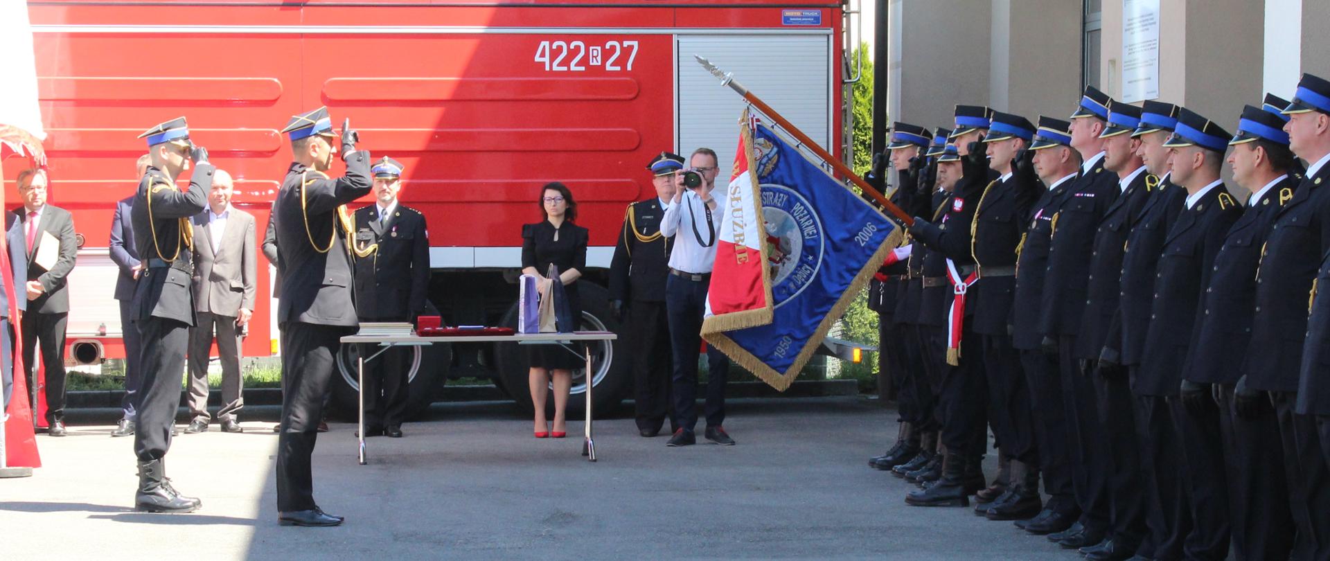 Zdjęcie wykonane na zewnątrz. Po prawej stronie stoją strażacy ubrani w mundury wyjściowe, ustawieni w szeregu. Jeden ze strażaków trzyma pochylony do przodu sztandar Komendy Powiatowej Państwowej Straży Pożarnej. Po lewej stronie zdjęcia dwóch oficerów Państwowej Straży Pożarnej salutuje sztandarowi. W tle stoją goście uczestniczący w uroczystości.
