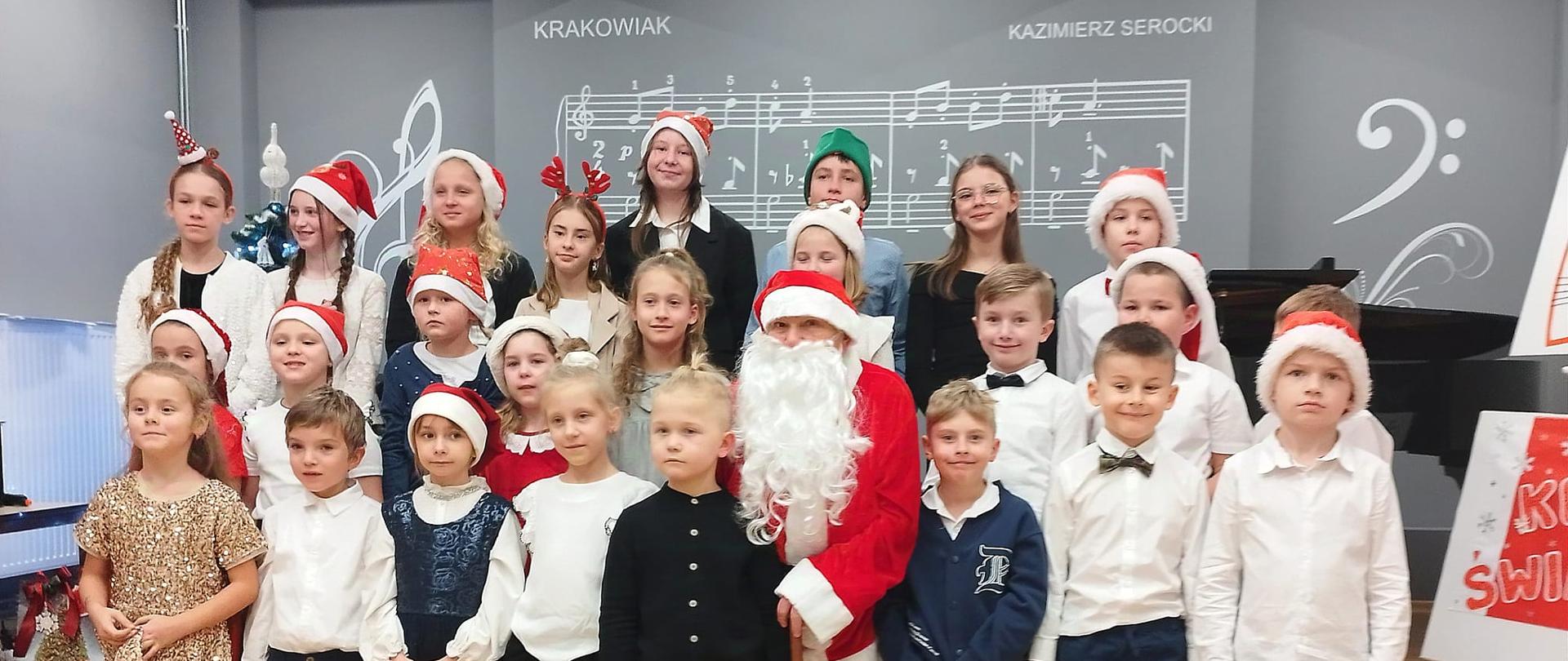 Na scenie w Szkole Muzycznej stoje uczniowie klas pierwszych ubrani odświętnie na głowach maja mikołajkowe czapki po środku stoi święty mikołaj