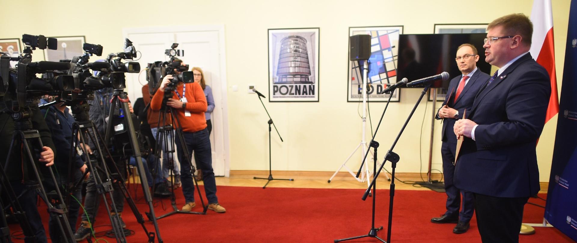 W sali wyłożonej czerwona wykładziną dywanową stoi z prawej wiceminister Rzymkowski i mówi do mikrofonu na stojaku, z lewej kilku dziennikarzy stojących przy kamerach na statywach.
