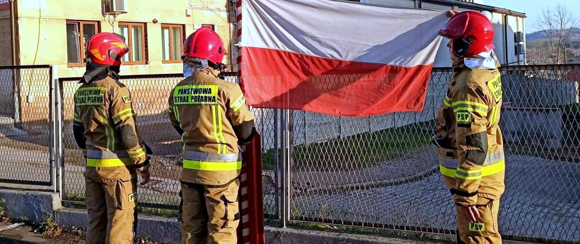 Na zdjęciu widać poczet flagowy wciągający flagę na maszt. Wszyscy strażacy ubrani są w umundurowanie specjalne, a na głowie mają hełmy.