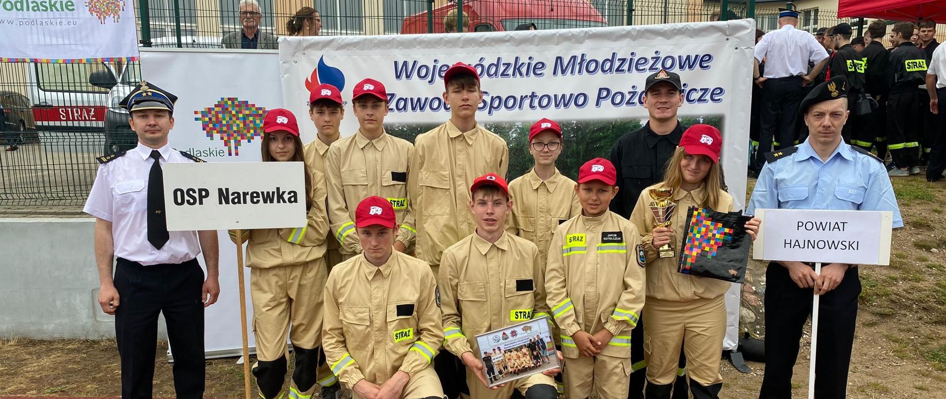 Na boisku w szeregu stoją członkowie Młodzieżowej Drużyny Pożarniczej. Po bokach strażak w białej i niebieskiej koszuli.