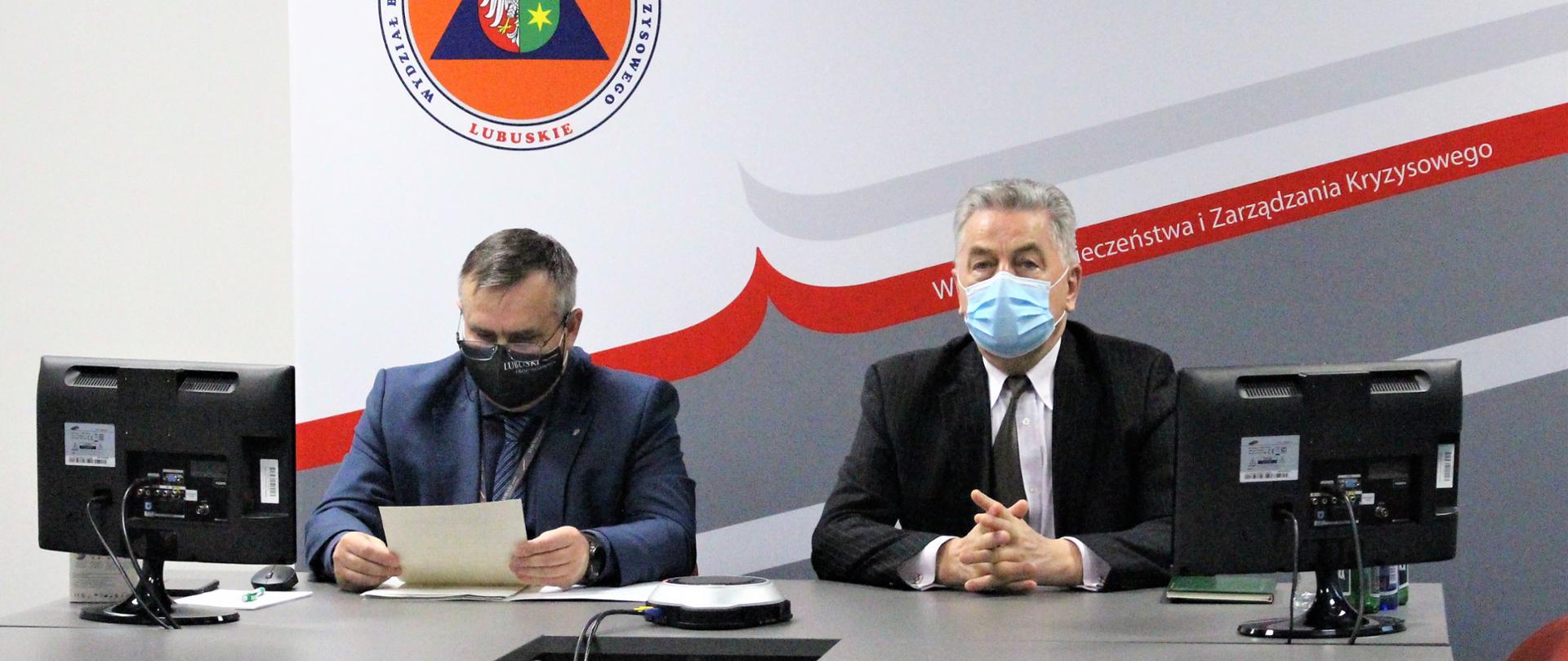 Wicewojewoda i dyrektor BZK siedzą obok siebie przy stole z dwoma ekranami po bokach. Za nimi stoi ścianka promocyjna urzędu z logo wydziału 