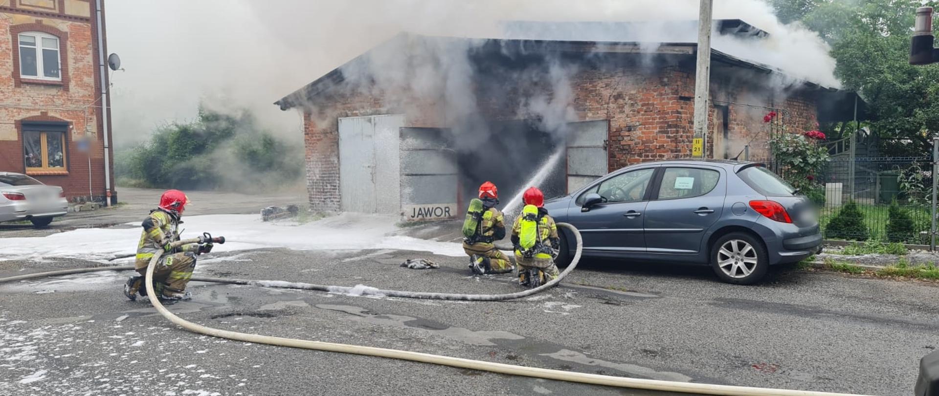 Obraz przedstawia strażaków gaszących pożar garażu