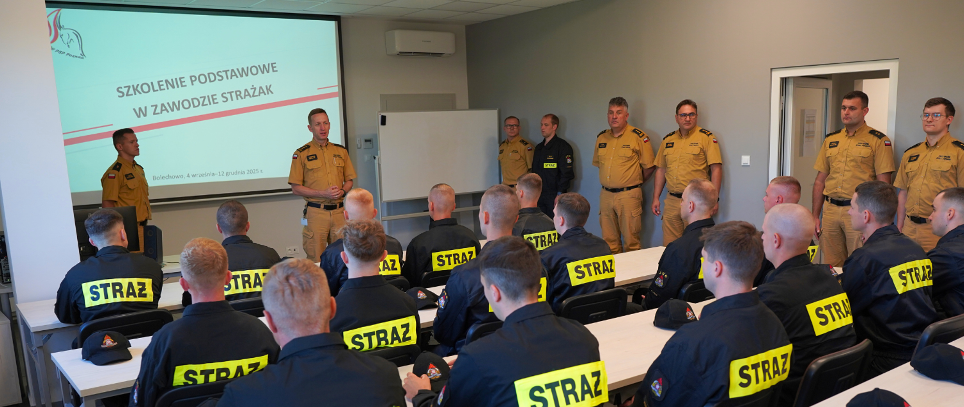 Pan Komendant stojąc na środku sali zabiera głos i wita siedzących w ławkach przed nim adeptów pożarnictwa, w sali.