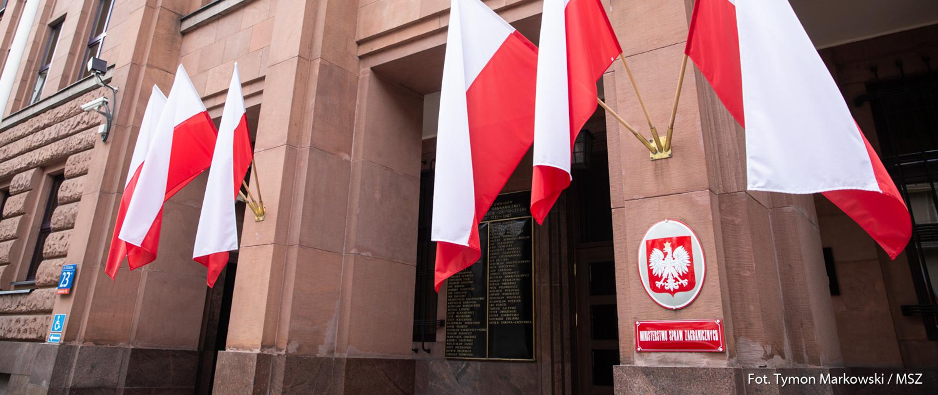 Front budynku Ministerstwa Spraw Zagranicznych, tabliczka z nazwą, godło i sześć biało-czerwonych flag.