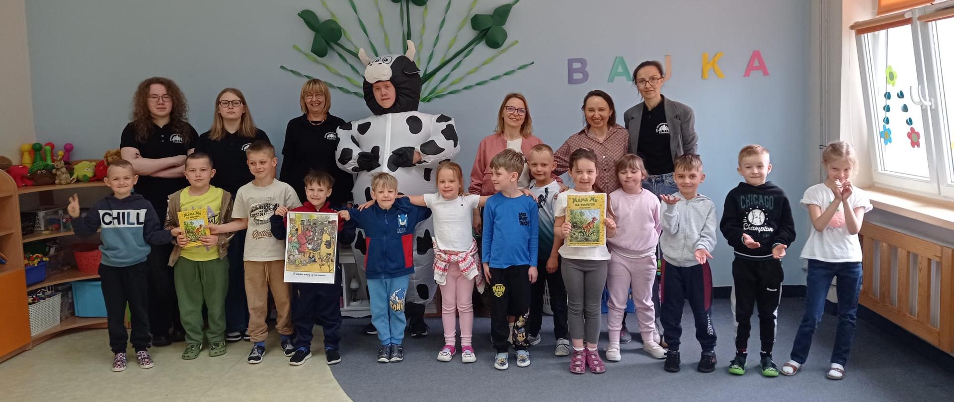 Grupa przedszkolaków, młodzież szkolna oraz nauczycielki pozują do zdjęcia w sali przedszkolnej. W środku stoi osoba w nadmuchiwanym stroju łaciatej krowy (Mama Mu).