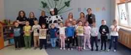 Grupa uśmiechniętych przedszkolaków, młodzież szkolna oraz nauczycielki pozują wspólnie do zdjęcia w kolorowej sali przedszkolnej. W centralnej części stoi osoba przebrana w duży, nadmuchiwany strój łaciatej krowy (Mama Mu), a dzieci trzymają w rękach książeczki oraz ilustracje.