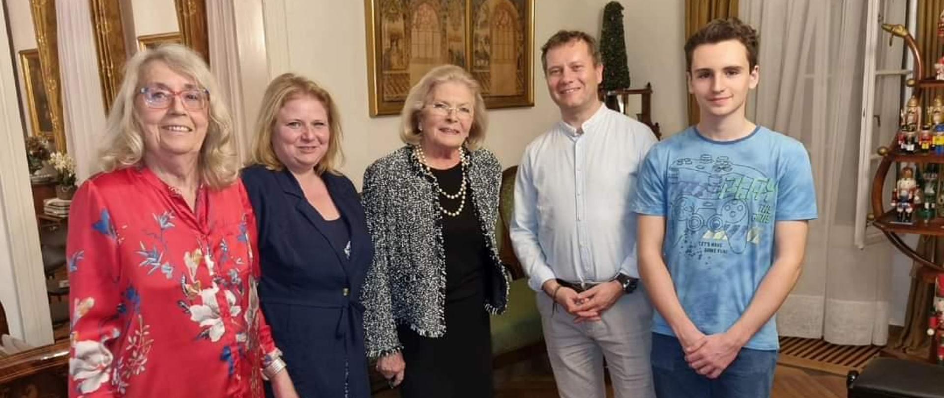 na zdjęciu widać od lewej strony kobietę w czerwonej kwiecistej bluzce, blond włosach i okularach , obok kobieta w blond włosach i granatowym stroju, obok kobieta blondynka w czarnym stroju na szyi ma załozone białe korale obok meżczyzna w jasnym ubraniu i blond włosach obok mężczyzna w niebieskiej koszulce i niebieskich spodniach. . Zdjęci w tle z ozdobnym wnetrzem na onie wiszą złote zasłony, na ścianie obraz w złotej ramie