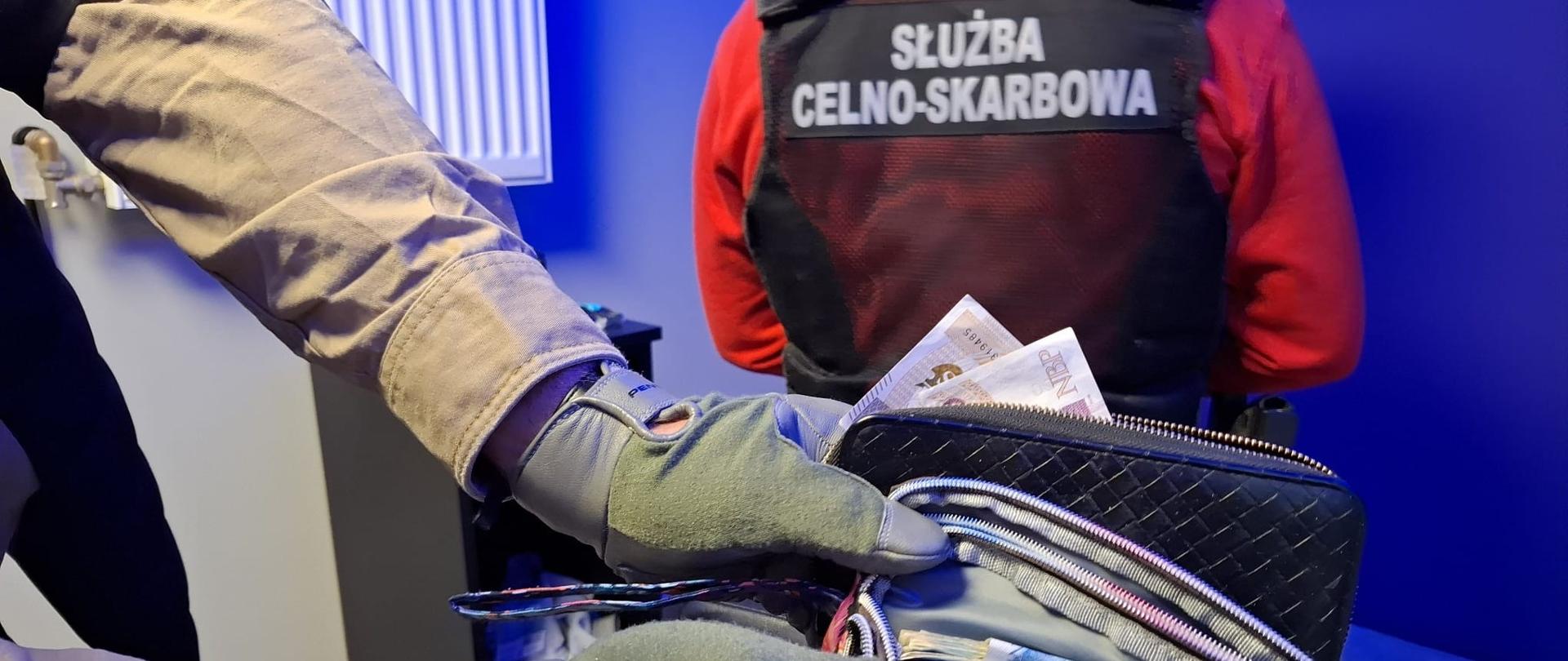 Funkcjonariusz w rękawicach przegląda saszetkę z banknotami. W tle widoczna jest kamizelka z napisem „Służba Celno-Skarbowa"