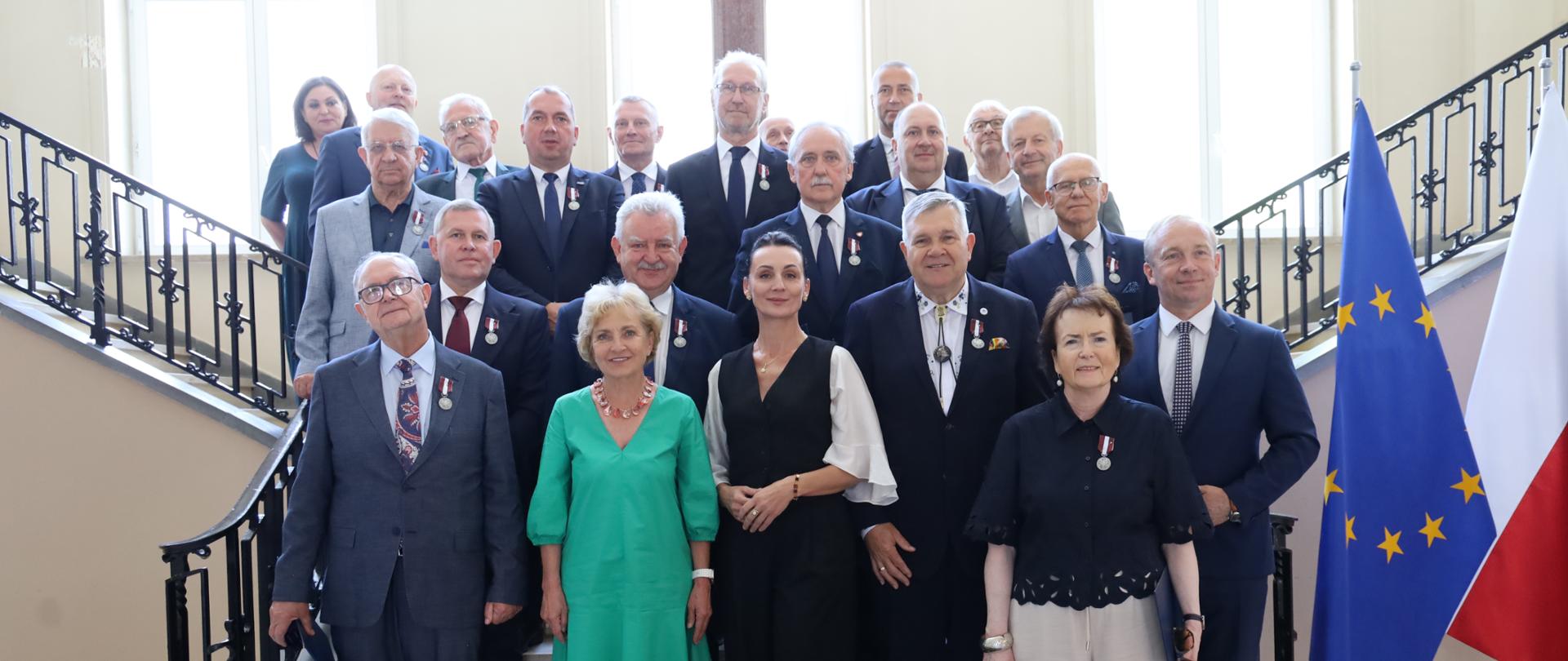Zdjęcie grupowe przedstawiające dwadzieścia osób pozujących na wewnętrznych, kamiennych schodach z ozdobną, czarną, metalową barierą po lewej stronie. Większość grupy stanowią mężczyźni w eleganckich garniturach, z których wielu ma przypięte do klap odznaczenia.