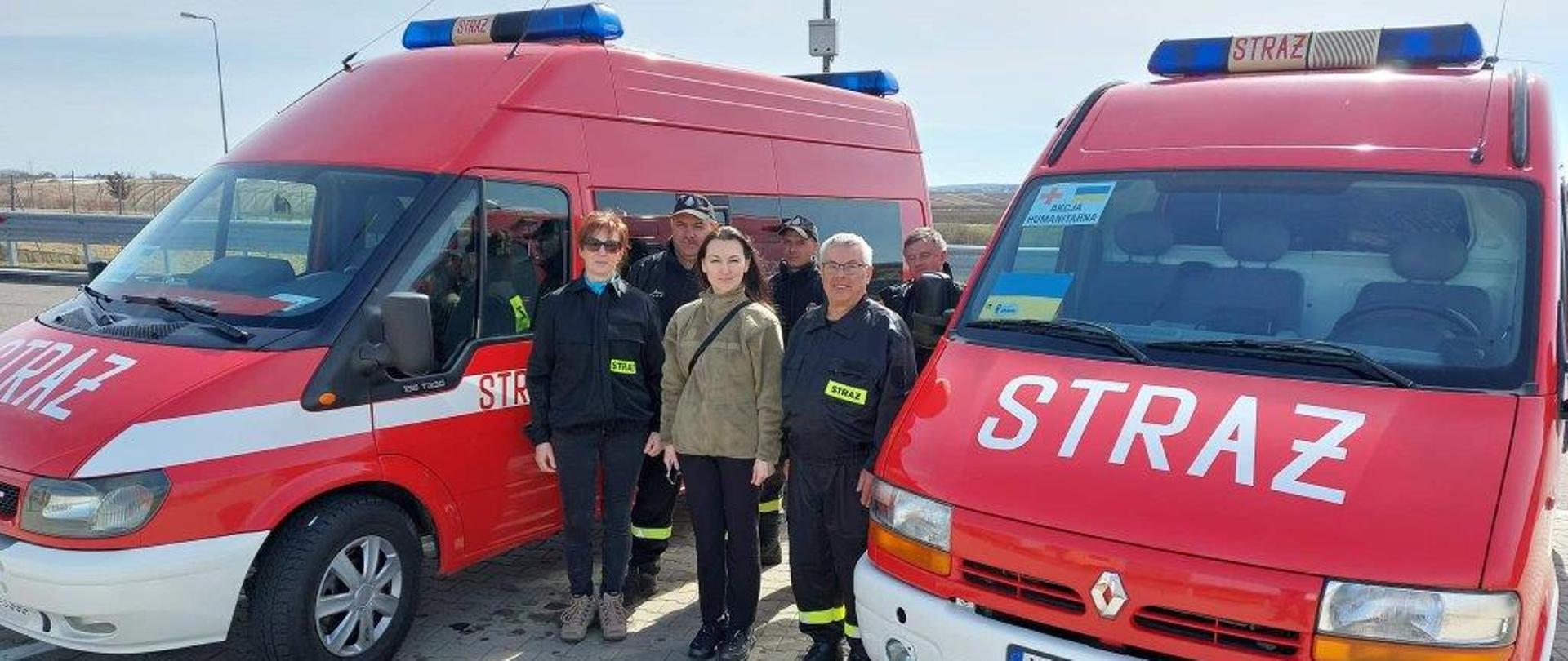 Dwa wozy strażackie z darami obok druhowie przewożący dary