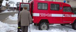 Na zdjęciu widać strażaka w czarnym ubraniu z napisem STRAŻ na plecach pomagającego starszej osobie wsiąść do osobowego terenowego samochodu strażackiego