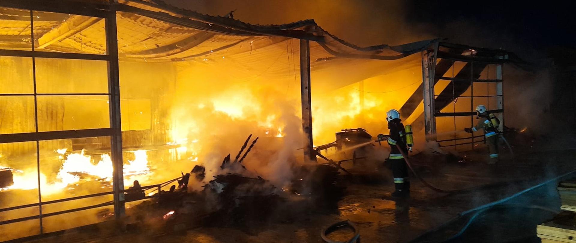 Strażacy podający prądy wody do objętej pożarem wiaty magazynowej