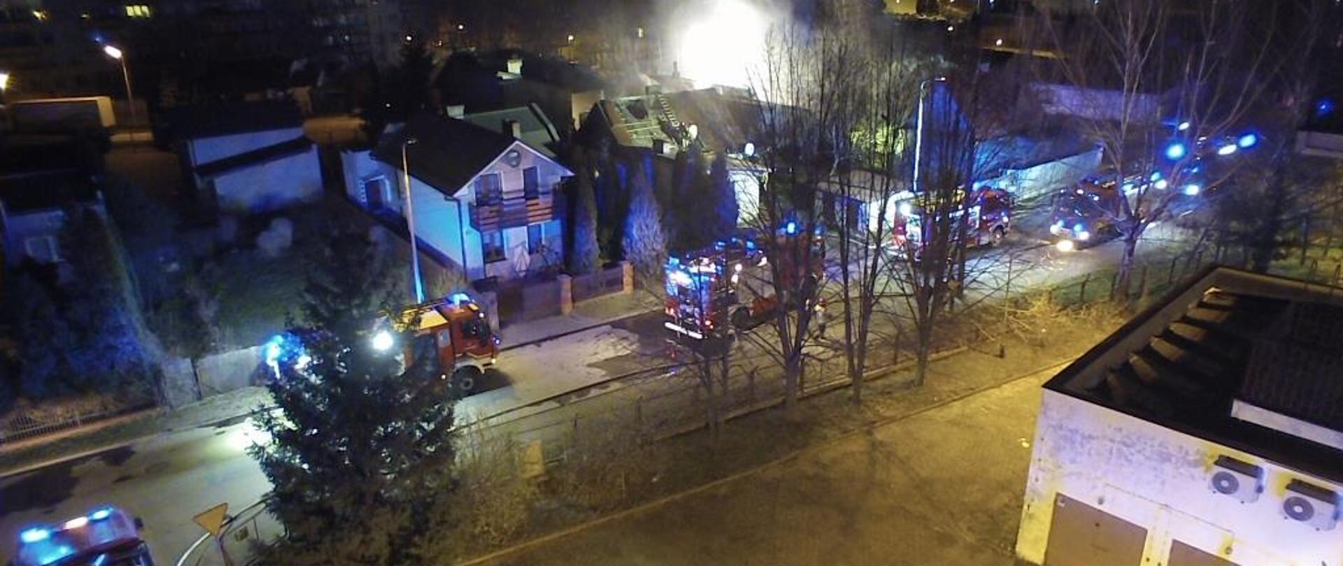 Zdjęcie wykonane w porze nocnej przy użyciu drona. Na zdjęciu widać zabudowę mieszkalną jednorodzinną. Przy ulicy pomiędzy domami ustawione są pojazdy ratowniczo – gaśnicze. Pojazdy w kolorze czerwonym mają włączoną niebieską świetlną sygnalizację alarmową. W oddali widać jak nad kilkoma budynkami unosi się dym.