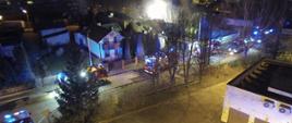 Zdjęcie wykonane w porze nocnej przy użyciu drona. Na zdjęciu widać zabudowę mieszkalną jednorodzinną. Przy ulicy pomiędzy domami ustawione są pojazdy ratowniczo – gaśnicze. Pojazdy w kolorze czerwonym mają włączoną niebieską świetlną sygnalizację alarmową. W oddali widać jak nad kilkoma budynkami unosi się dym.
