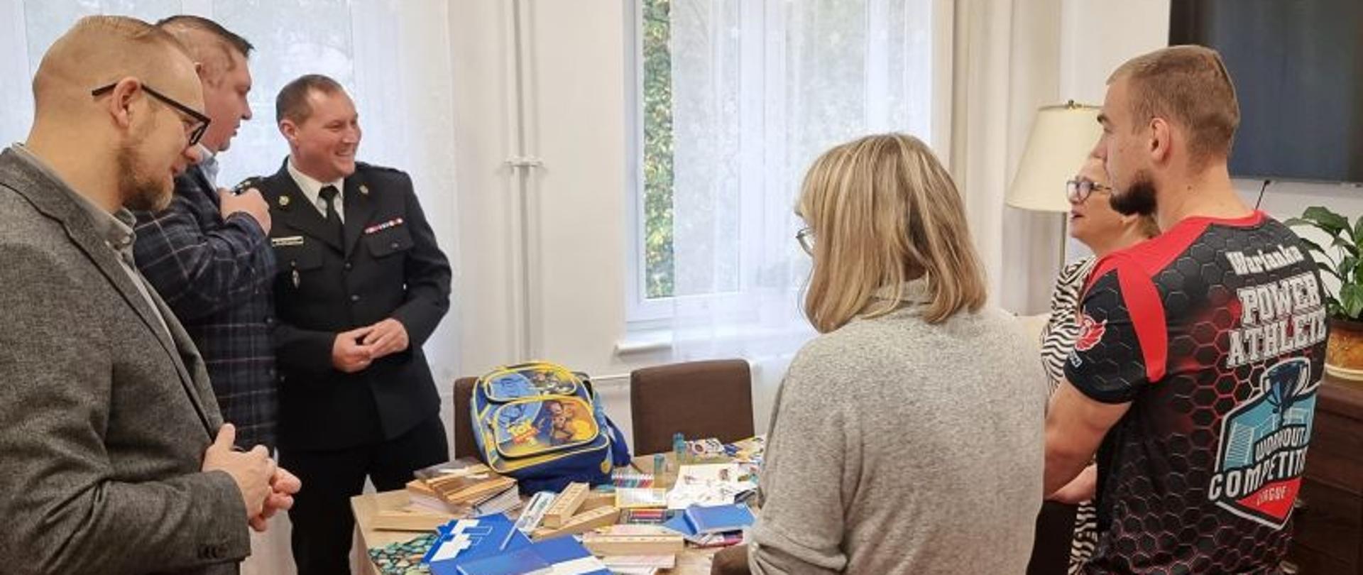 Zdjęcie przedstawia 4 mężczyzn i 2 kobiety. Wszyscy stoją przy stole, na którym leżą materiały piśmiennicze i biurowe. W tle okno zasłonięte firanką. Wśród mężczyzn Komendanta w galowym mundurze. Po prawej stronie na ścianie wisi telewizor.