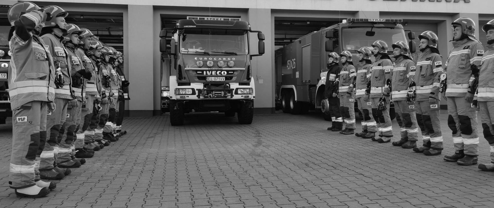 strażacy ustawieni w szeregach na przeciwko siebie stoją na baczność ubrani w stroje specjalne za nimi ustawione są samochody ratowniczo gaśnicze w tle znajduje się budynek Państwowej Straży Pożarnej