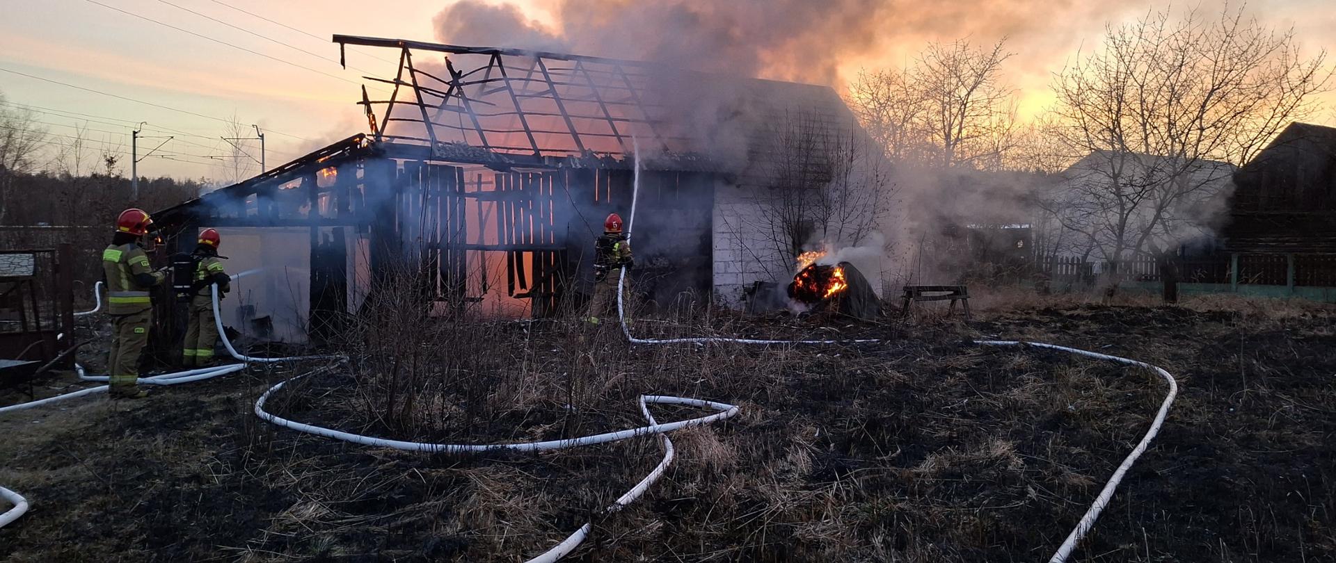 Pożar budynku gospodarczego o konstrukcji częściowo drewnianej, częściowo murowanej. W budynku widać płomienie a nad budynkiem unosi się gęsty dym. Strażacy podają dwa prądy wody w natarciu do wnętrza budynku.