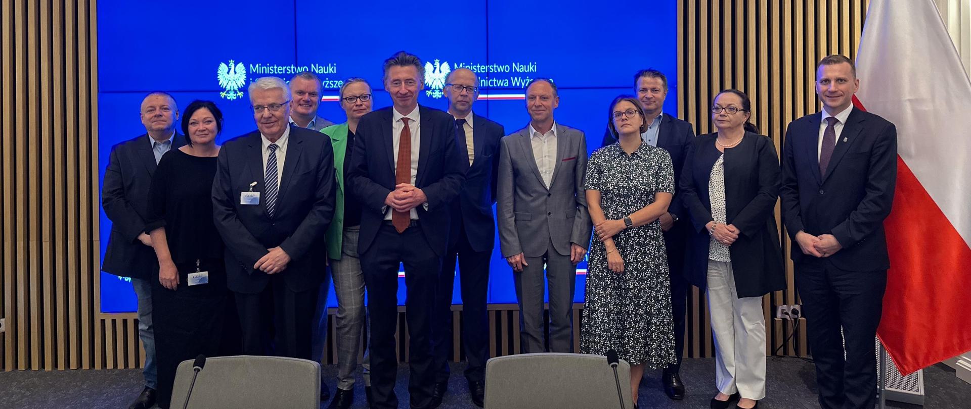 Zdjęcie zbiorowe, pod wbudowanym w ścianę wielkim ekranem stoi grupa dwunastu osób, pośrodku wiceminister Gdula, z boku z tyłu polska flaga.