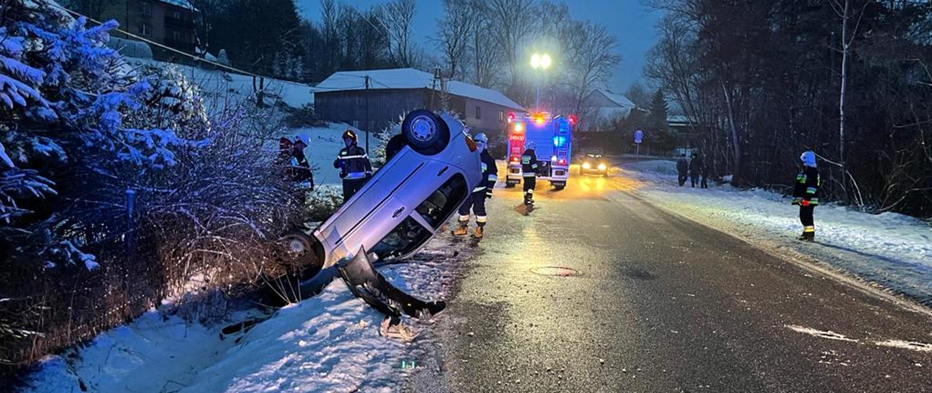 Samochód osobowy na dachu poza drogą. Obok strażacy oraz samochód pożarniczy.