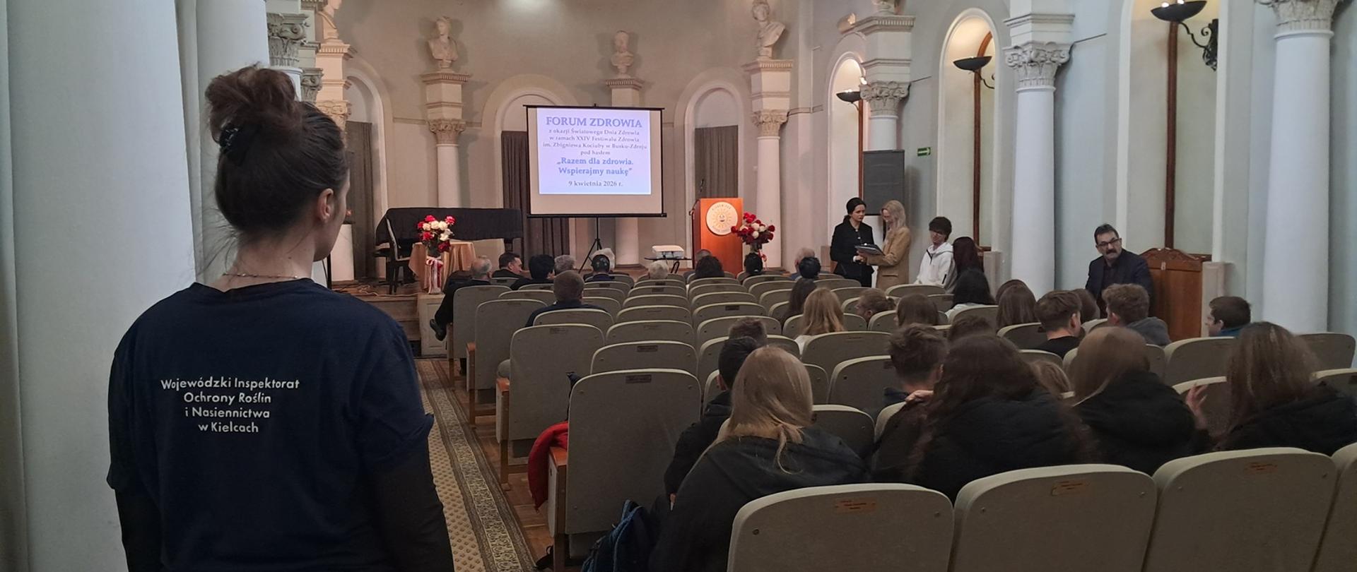 Wnętrze sali wykładowej z ozdobnymi kolumnami. Pośrodku sali znajdują się rzędy jasnych krzeseł, na których siedzą ludzie, głównie młodzież. Część osób rozmawia ze sobą a inne patrzą w kierunku sceny. Na środku sceny stoi ekran projekcyjny, na którym widać napis „FORUM ZDROWIA z okazji Światowego Dnia Zdrowia w ramach XXIV Festiwalu Zdrowia im. Zbigniewa Kociuby w Busku-Zdroju pod hasłem ”Razem dla zdrowia wspierajmy naukę” 9 kwietnia 2026r.” Po lewej stronie sali przodem do sceny stoi pracownik inspekcji w granatowej koszulce z napisem Wojewódzki Inspektorat Ochrony Roślin i Nasiennictwa w Kielcach.