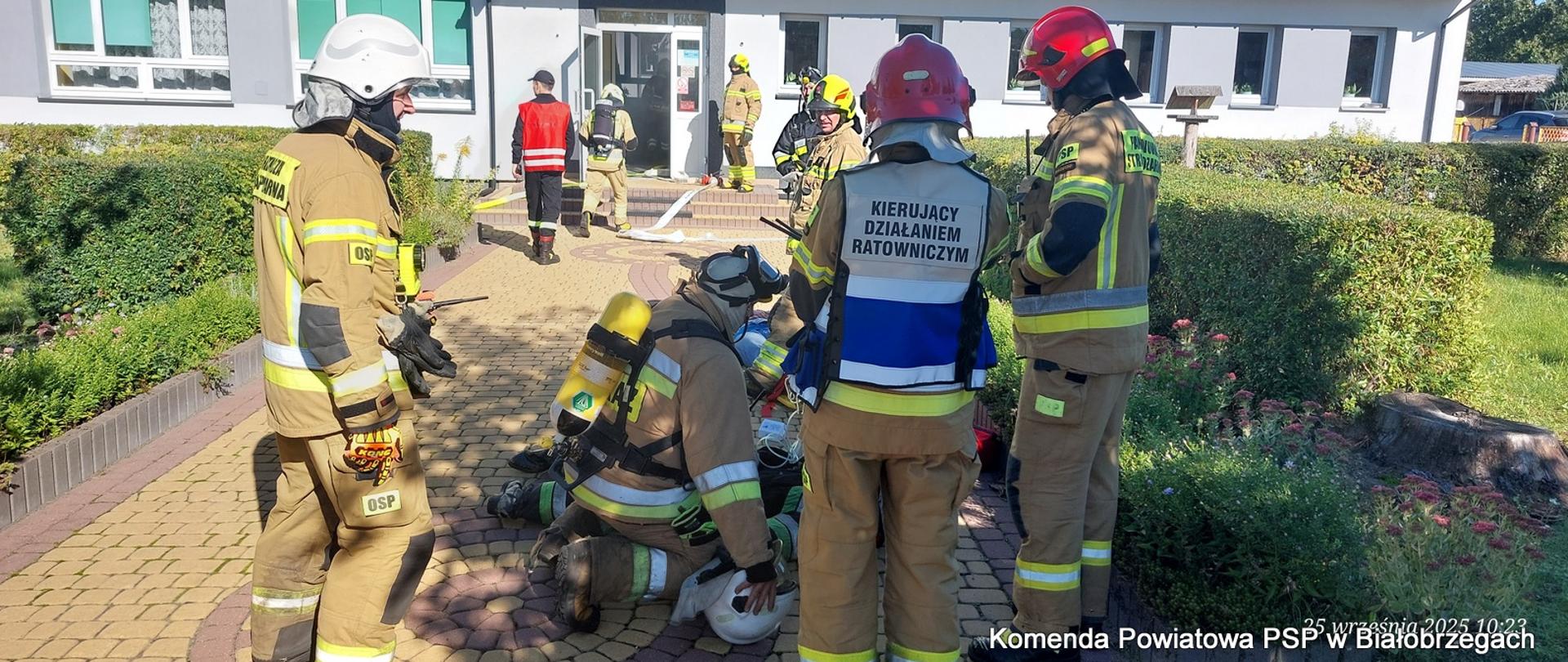 Grupa strażaków stoi na chodniku przed budynkiem. Jeden z nich leży na ziemi i jest opatrywany przez pozostałych. Na plecach jednego ze strażaków widać napis "KIERUJĄCY DZIAŁANIEM RATOWNICZYM".