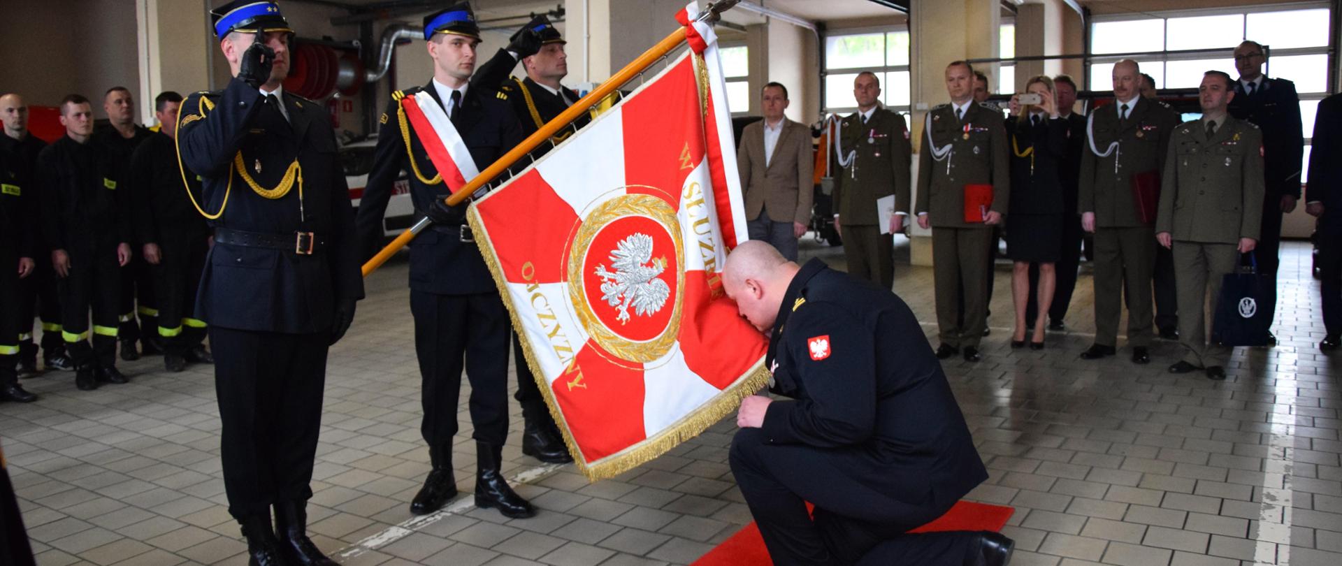 Komendant Powiatowy PSP w Mińsku Mazowieckim klęka przed sztandarem KP PSP w Mińsku Mazowieckim symbolicznie go całując, w tle zebrani goście oraz pracownicy i strażacy 