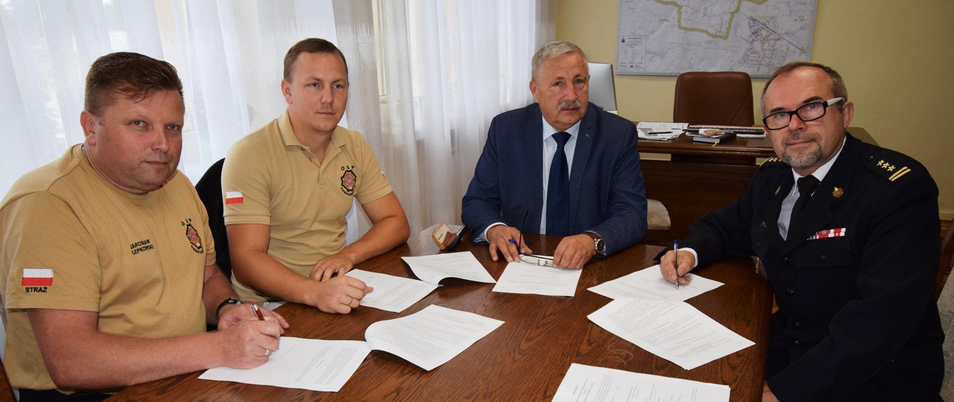 Cztery osoby siedzą za stołem i podpisują porozumienie. Z lewej dwóch strażaków OSP w koszulkach z logo straży pożarnej, na środku osoba w garniturze, z prawej oficer Państwowej Straży Pożarnej w mundurze wyjściowym. W tle, z lewej strony okna z firankami, z prawej na ścianie wisi mapa.