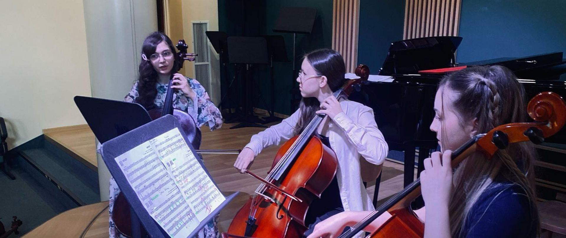 Na zdjęciu widać jak 3 uczennice grają na wiolonczelach przed nimi stoją 2 pulpity na których stoją nuty w tle widoczna Zielona ściana i fortepian