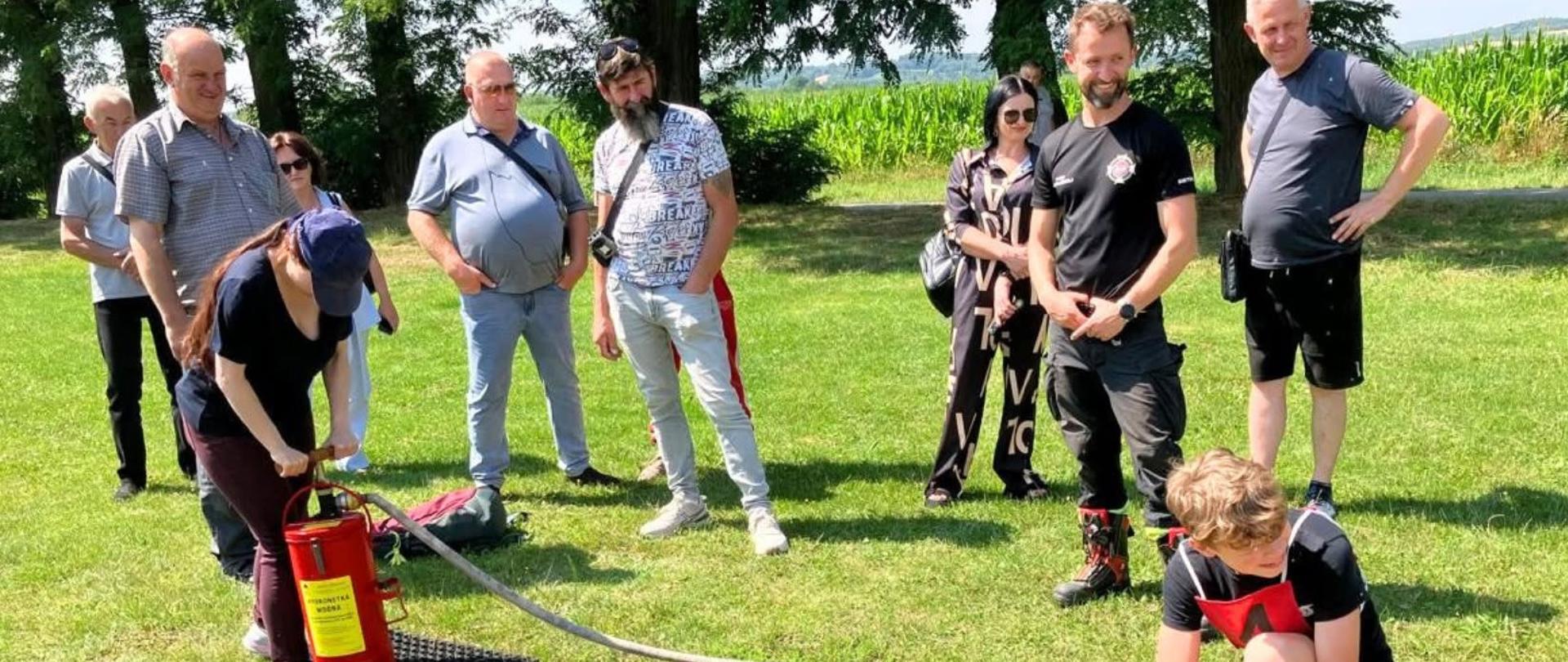 Na trawniku stoi grupa osób i patrzy na dwoje nastolatków testujących sprzęt strażacki 