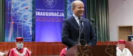 Wojewoda Mazowiecki przemawiający do mikrofonu w trakcie uroczystości. Za Wojewodą siedzący profesorowie uczelni ubrani w togi. 