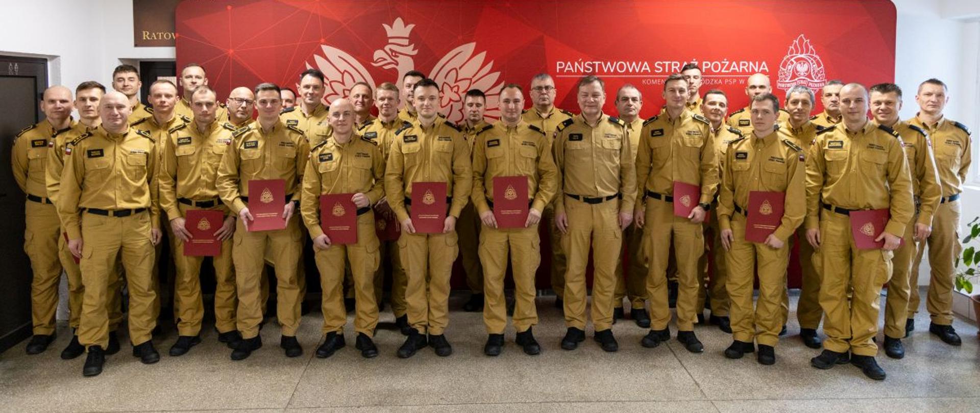 Grupa funkcjonariuszy PSP stoi w szeregu, część z nich trzyma czerwone foldery z wyróżnieniami; tłem jest ścianka Komendy Wojewódzkiej PSP w Opolu, co podkreśla uroczysty charakter wydarzenia.