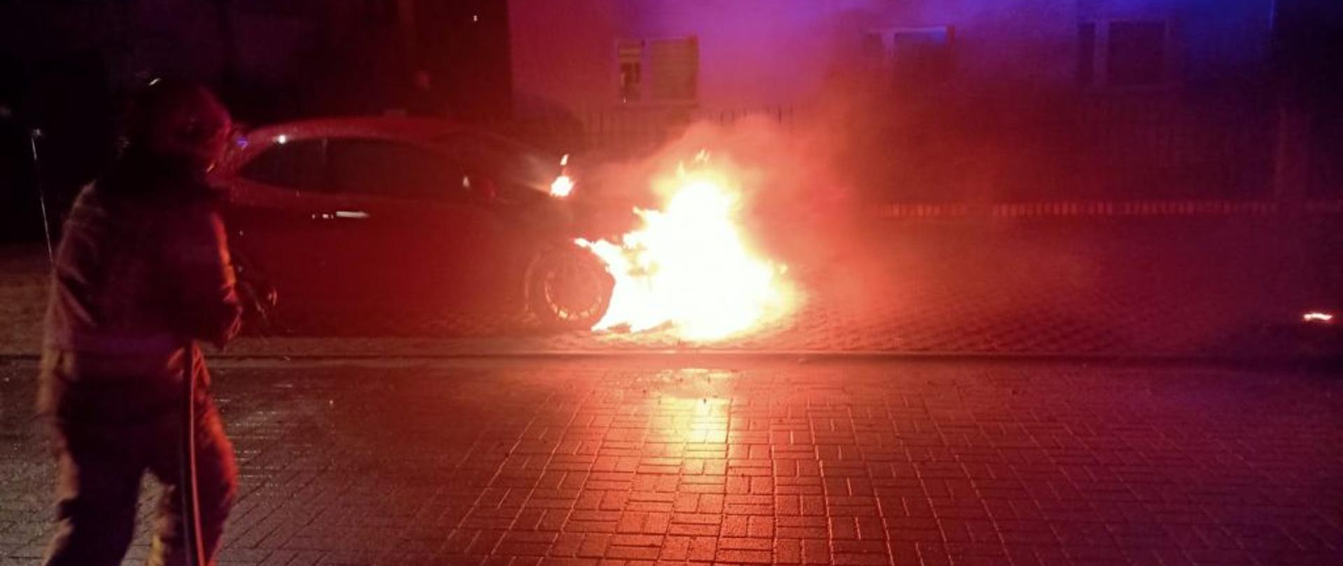 Widok na palące się auto. Zdjęcie wykonane w nocy. pożarem objęty jest przód auta. Po lewej stronie zdjęcia stoi strażak w ubraniu bojowy trzymający prądownicę.