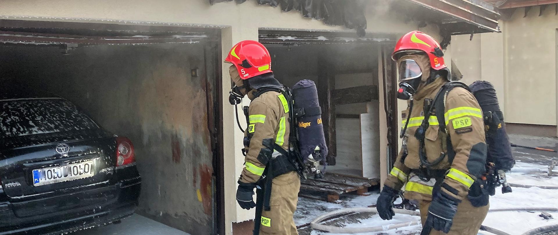 Dwóch strażaków w aparatach ODO i hełmach wchodzi do zadymionego garażu