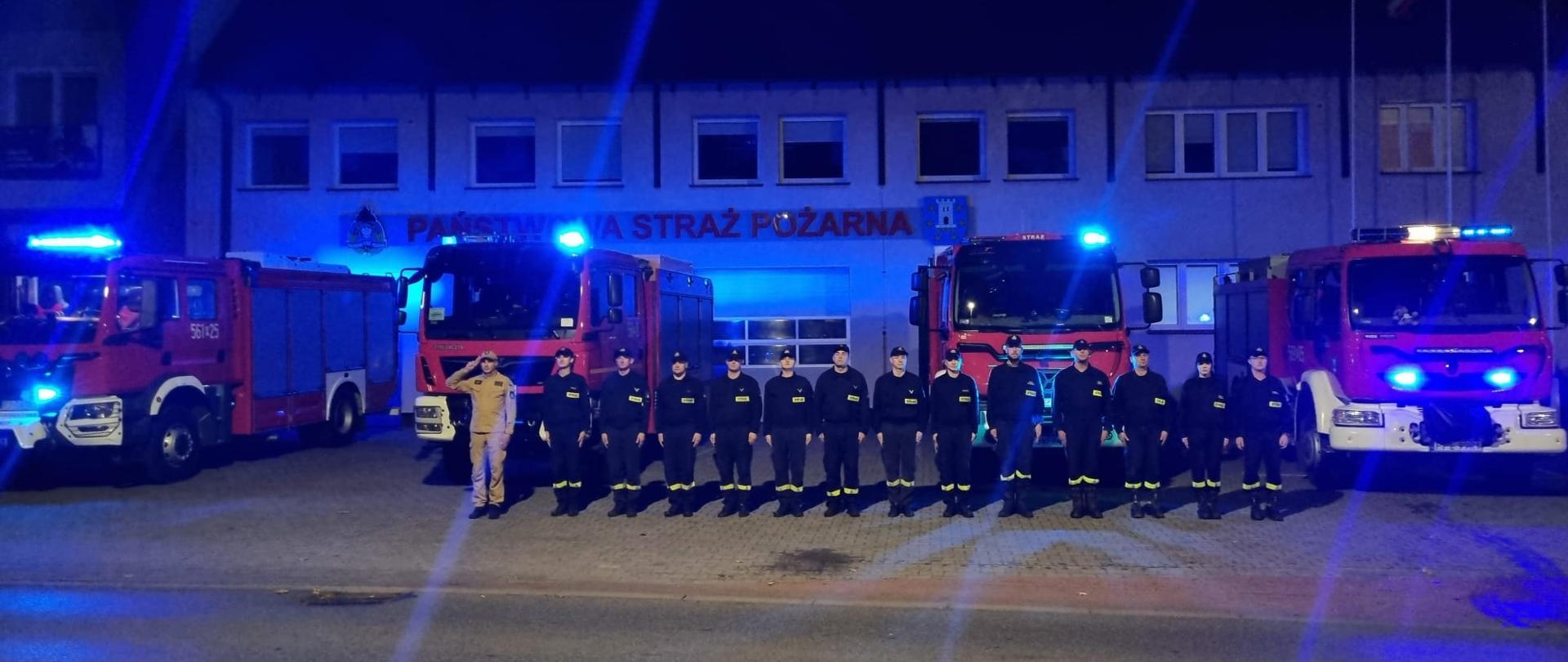 przed remizą strażacka stoją pojazdy z włączonymi sygnałami świetlnymi, przed autami stoją na baczność strażacy, wieczór