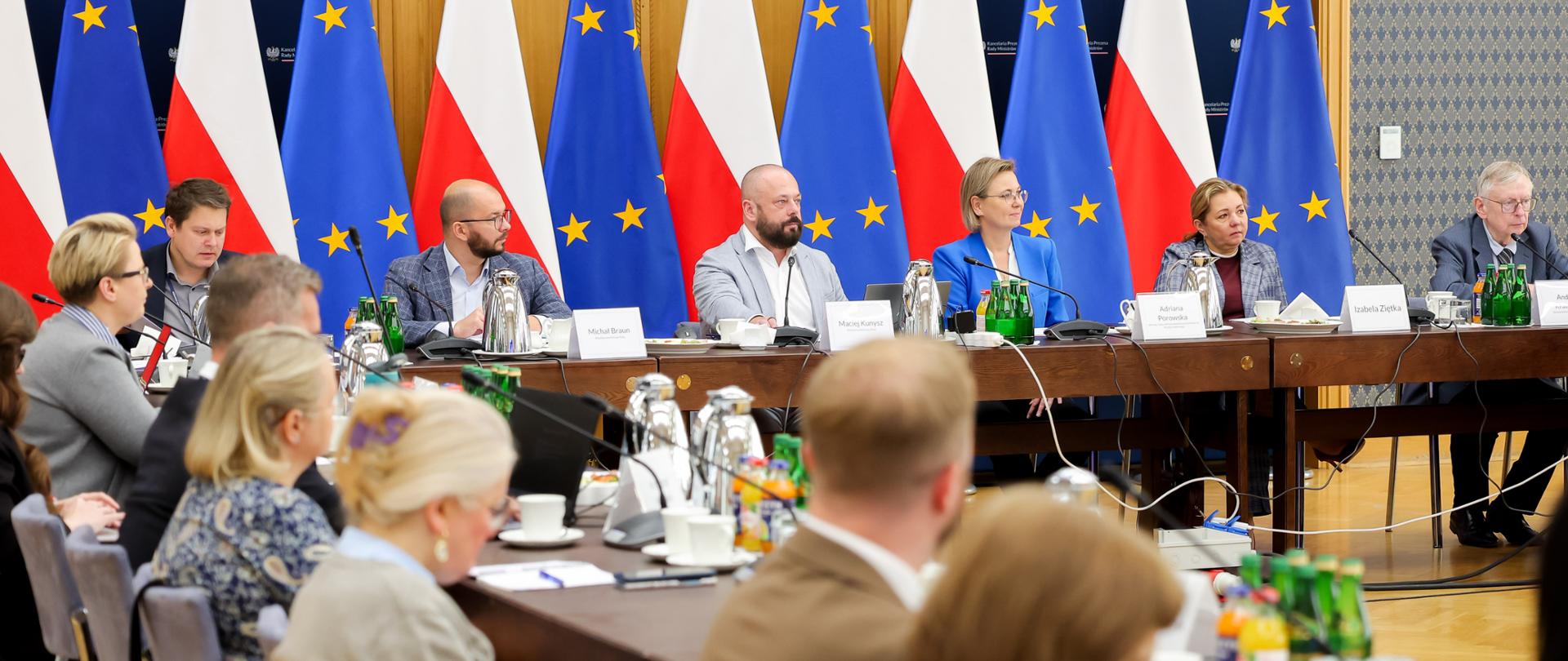 Trwa posiedzenie Rady Działalności Pożytku Publicznego. Na zdjęciu uczestnicy posiedzenia, siedzą przy stołach konferencyjnych. Na pierwszym planie widoczne są sylwetki uczestników. U szczytu stołu, na dalszym planie, widoczni między innymi Pani Adriana Porowska – Sekretarz Stanu w KPRM i Wiceprzewodnicząca Komitetu do spraw Pożytku Publicznego, Pan Łukasz Jachimowicz – Dyrektor Departamentu Społeczeństwa Obywatelskiego w KPRM oraz Pan Michał Braun – Dyrektor Narodowego Instytutu Wolności – Centrum Rozwoju Społeczeństwa Obywatelskiego. W tle flagi Polski i Unii Europejskiej.