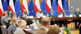 Trwa posiedzenie Rady Działalności Pożytku Publicznego. Na zdjęciu uczestnicy posiedzenia, siedzą przy stołach konferencyjnych. Na pierwszym planie widoczne są sylwetki uczestników. U szczytu stołu, na dalszym planie, widoczni między innymi Pani Adriana Porowska – Sekretarz Stanu w KPRM i Wiceprzewodnicząca Komitetu do spraw Pożytku Publicznego, Pan Łukasz Jachimowicz – Dyrektor Departamentu Społeczeństwa Obywatelskiego w KPRM oraz Pan Michał Braun – Dyrektor Narodowego Instytutu Wolności – Centrum Rozwoju Społeczeństwa Obywatelskiego. W tle flagi Polski i Unii Europejskiej.